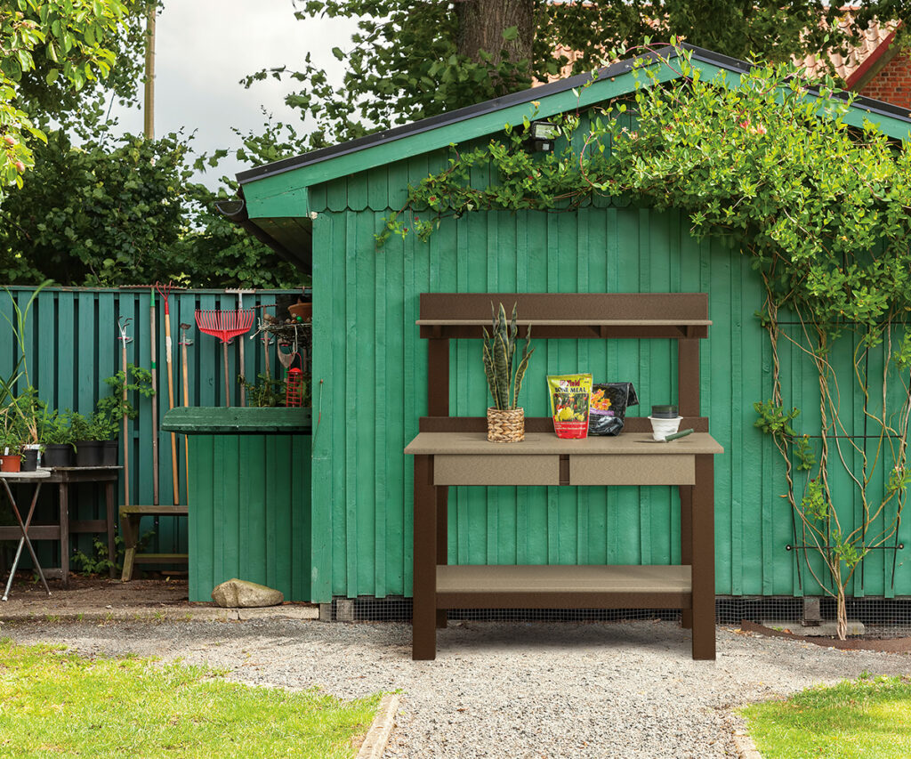 Outdoor potting bench with shelf beside green garden shed and climbing vines.
